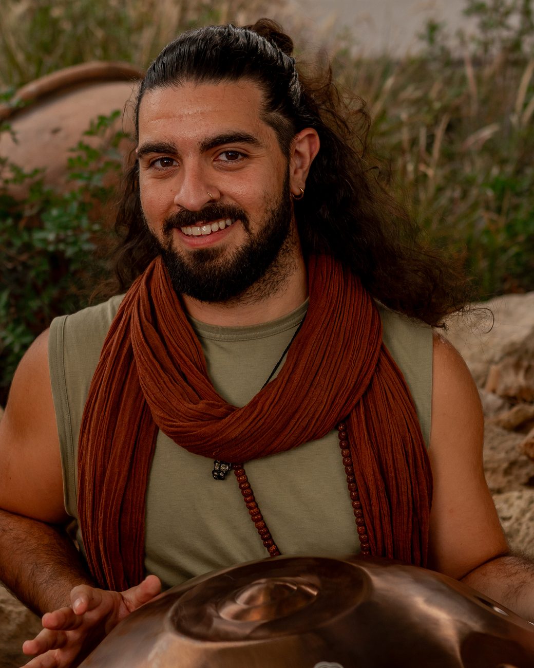 Emre Veçhi with a copper handpan among stones and tall grasses